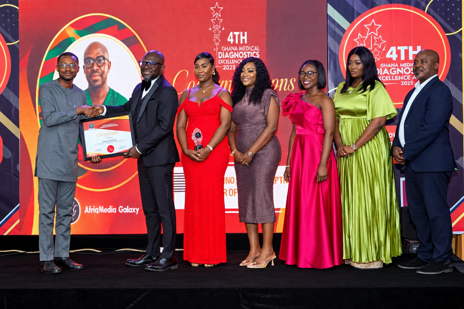 Representatives of Elixir Clinic on stage at the 4th Ghana Medical and Diagnostics Excellence Awards 2025, receiving a framed certificate and crystal award in front of the event's red backdrop.