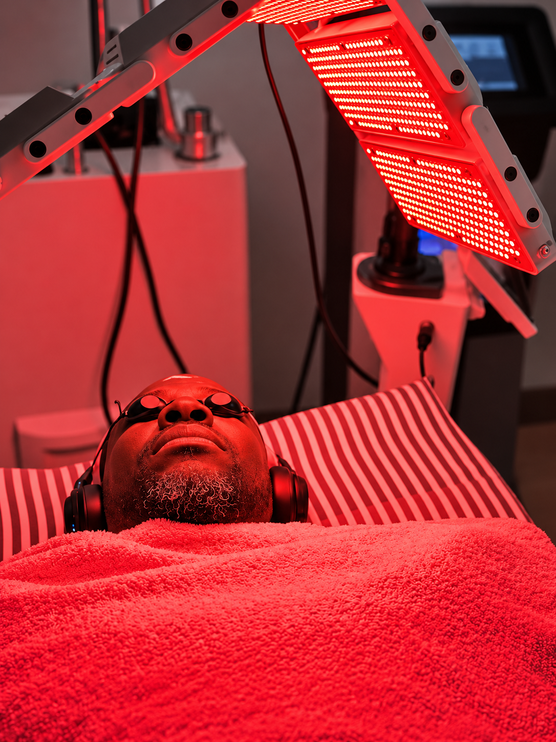 A patient reclining on a treatment bed at Elixir Clinic wearing black protective goggles and headphones, beneath a multi-panel red light array held on an articulated arm.
