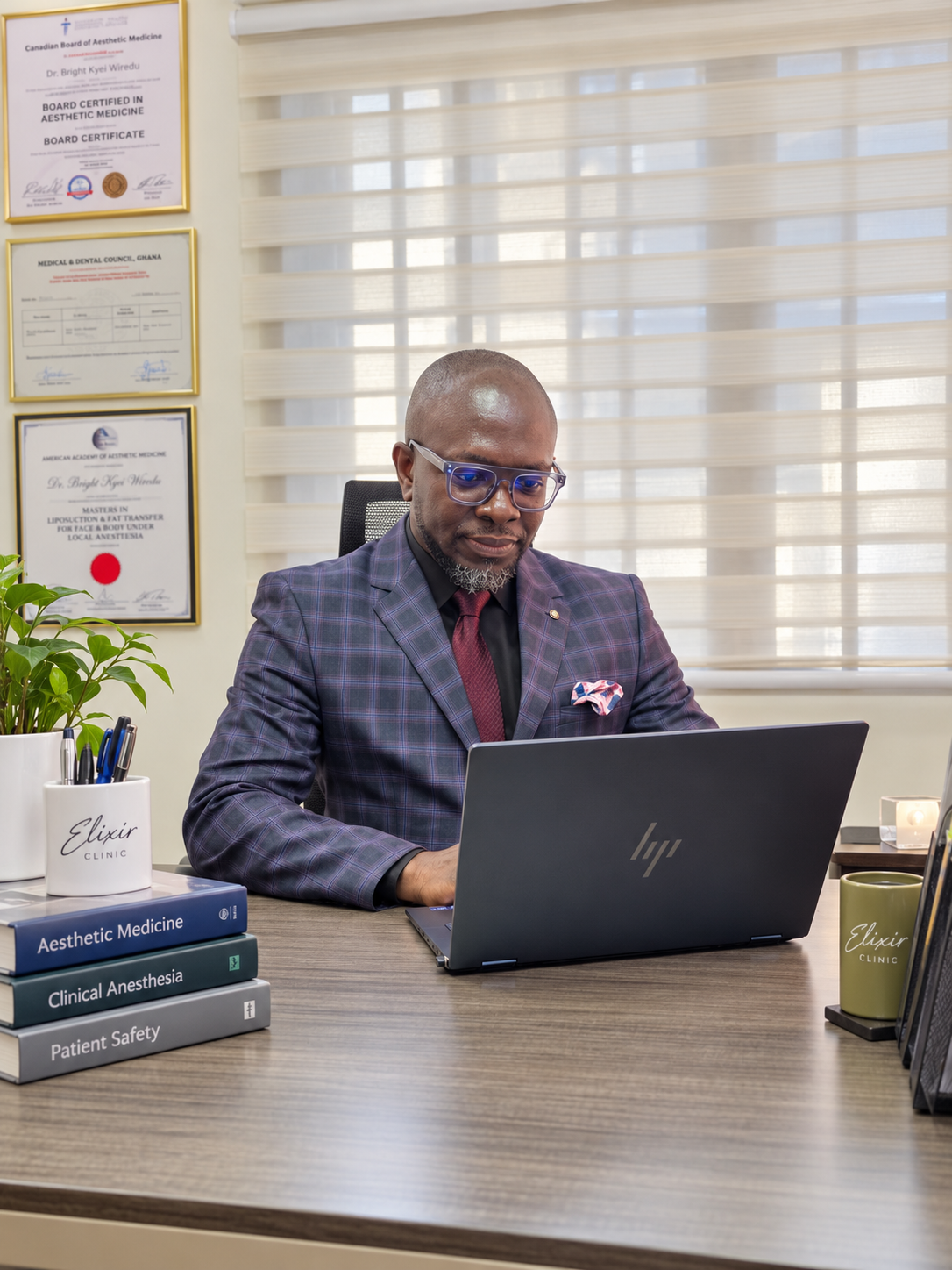 Dr. Bright Kyei Wiredu, Medical Director of Elixir Clinic, working at his desk in the consulting room in a checked blazer, with his framed certificates on the wall behind him.