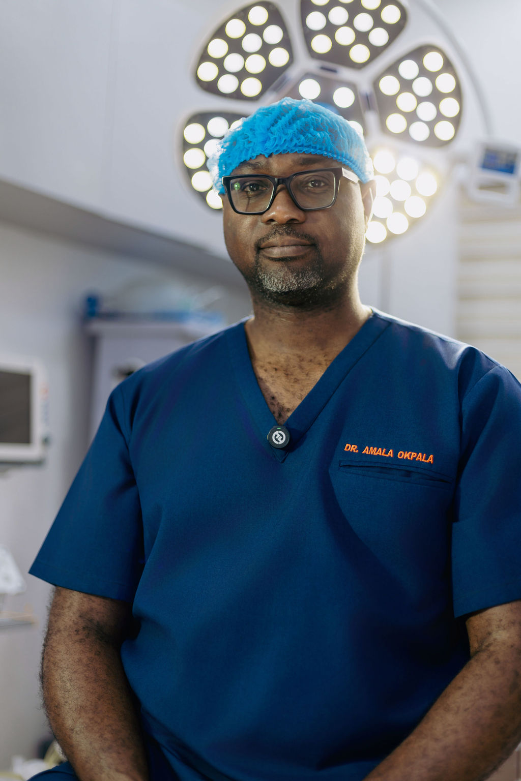 Dr Amalachukwu Okpala, Consultant Oncoplastic Breast Surgeon, standing in blue scrubs and a blue surgical cap beneath operating theatre lights, with “Dr. Amala Okpala” embroidered in orange on his scrub top.
