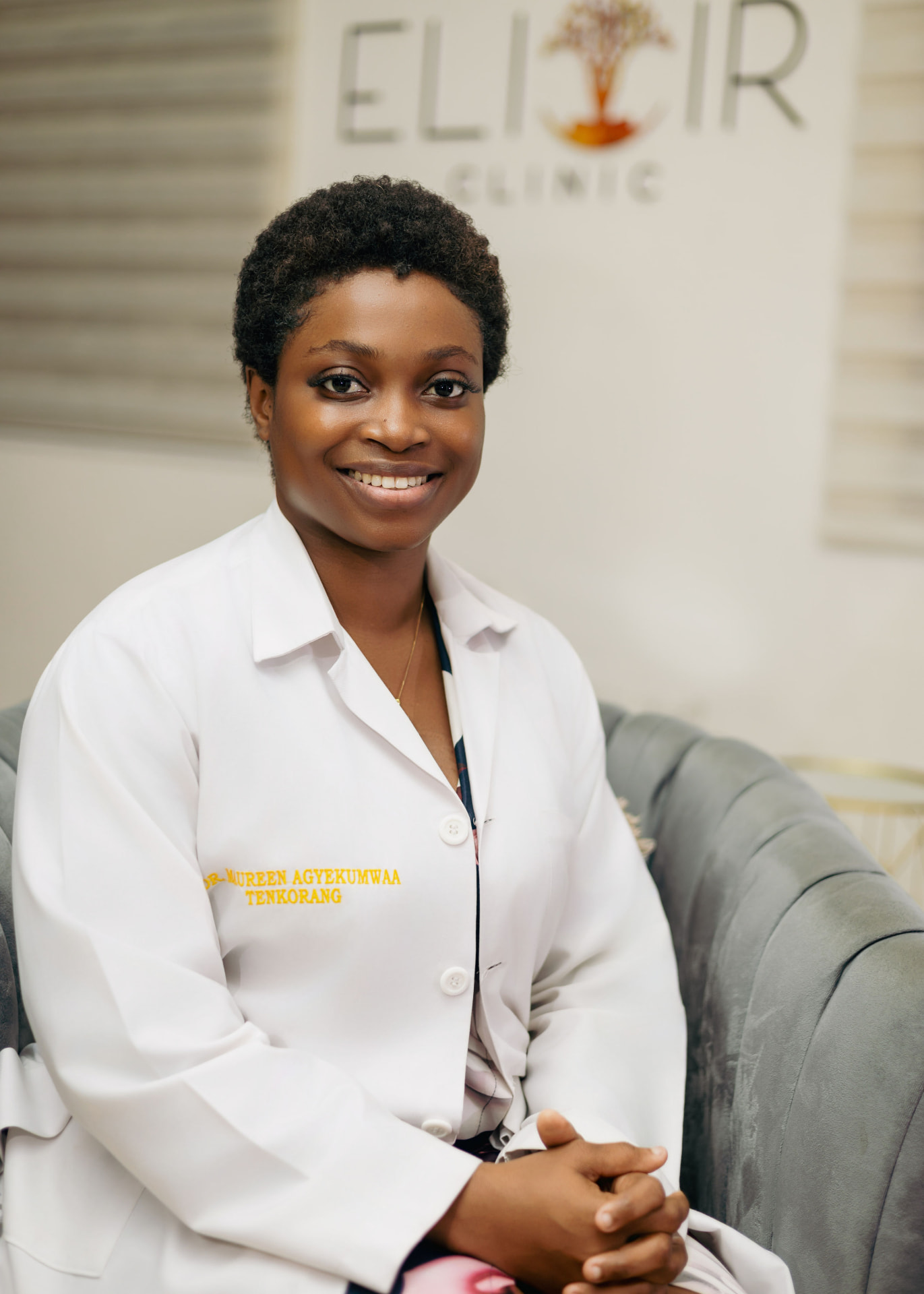 Dr. Maureen Agyekumwaa Tenkorang, Aesthetic Physician at Elixir Clinic, seated in a grey armchair in a white consultation coat embroidered with her name, with the clinic's Elixir signage on the wall behind her.