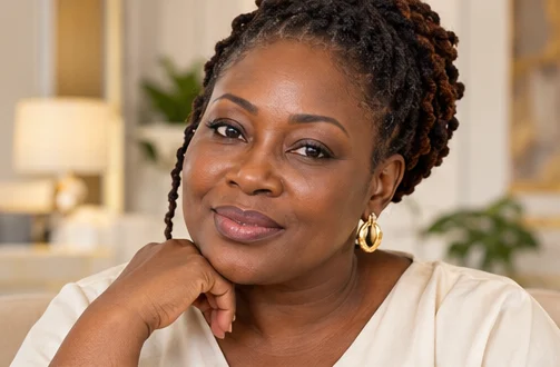 Smiling Ghanaian woman in her forties with natural skin texture, resting her chin on her hand in a bright clinic lounge.