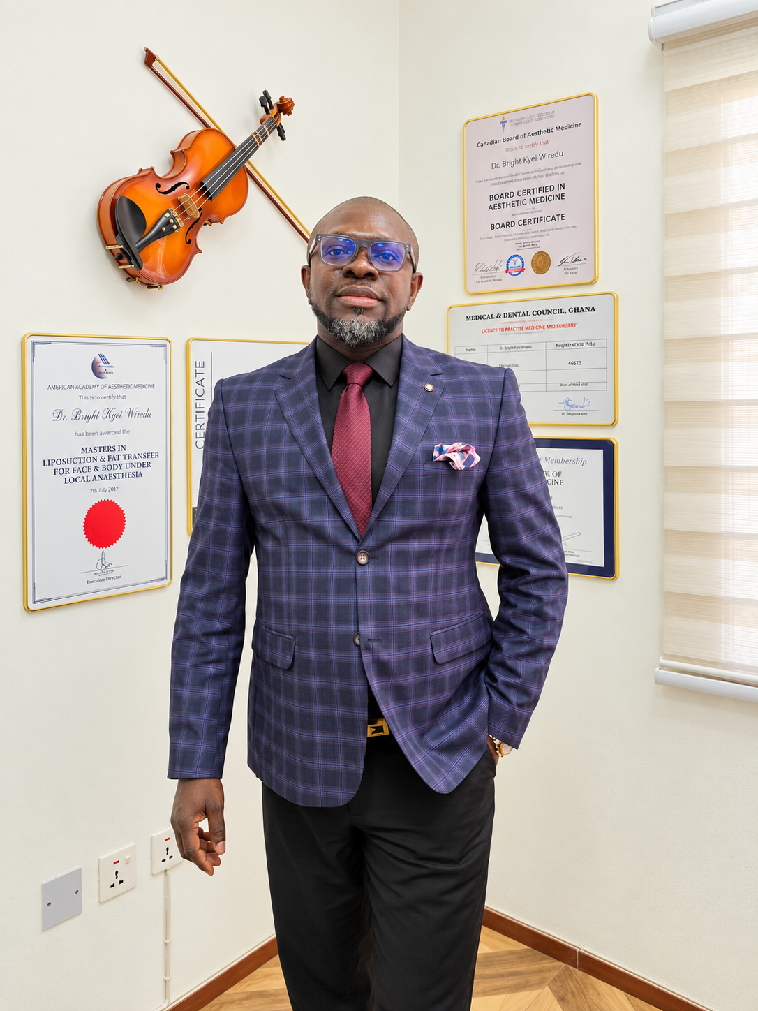 Dr. Bright Kyei Wiredu, Medical Director of Elixir Clinic, standing in his consultation room in a dark blazer and tie, with framed certificates on the wall behind him.