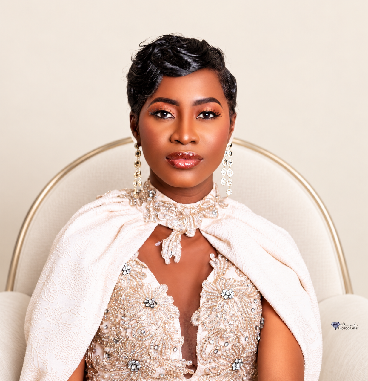 Portrait photograph of a woman in a cream beaded gown with crystal drop earrings and a choker, seated upright in a light armchair against a warm ivory studio backdrop.